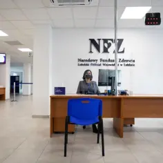 Pracownica NFZ za ladą w biurze, z logo NFZ na ścianie.